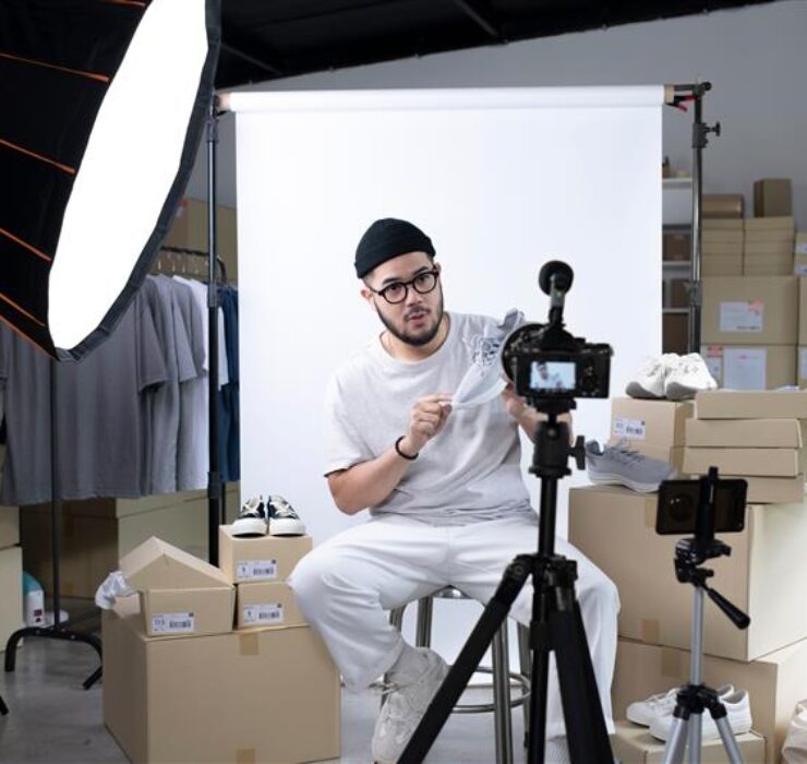 Influencer wearing a beanie and casual outfit, sitting in a studio setup with boxes and clothing in the background. He is recording content using a professional camera on a tripod under bright studio lights.