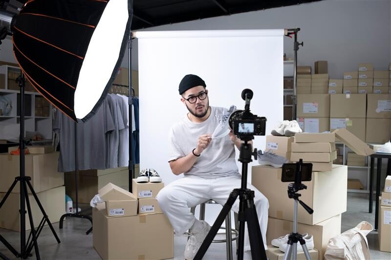 Influencer wearing a beanie and casual outfit, sitting in a studio setup with boxes and clothing in the background. He is recording content using a professional camera on a tripod under bright studio lights.